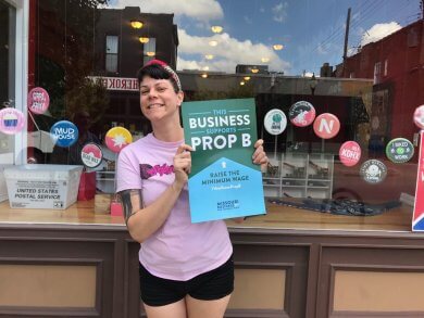 A woman holding up a sign that reads 'This Business Supports Prop B, Raise The Minimum Wage'